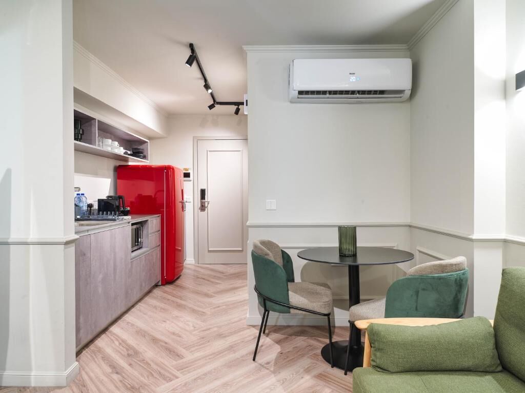 A compact kitchen with a red refrigerator, a dining area with two chairs and a small table, and a white wall-mounted air conditioning unit.