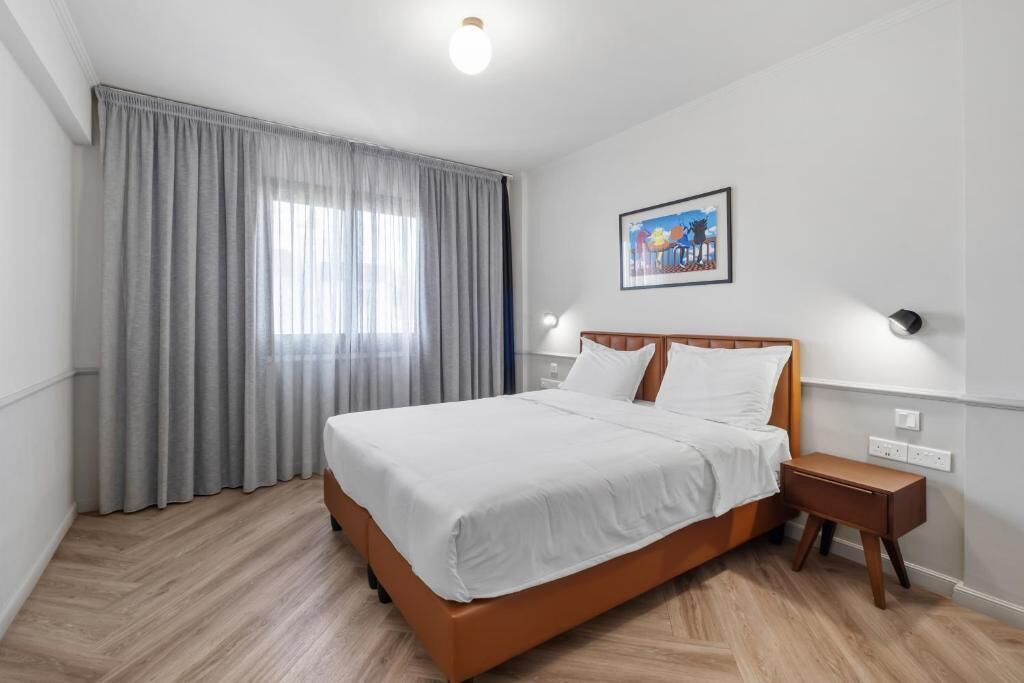 Une chambre d’hôtel avec un lit, une table de chevet, une lampe fixée au mur, une fenêtre avec des rideaux et un tableau accroché au mur.