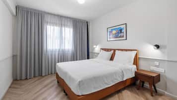 A hotel room with a bed, a nightstand, a wall-mounted lamp, a window with curtains, and a painting on the wall.