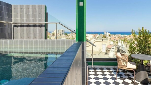 A rooftop pool with a checkered floor, a green pergola, and a view of the city and sea.