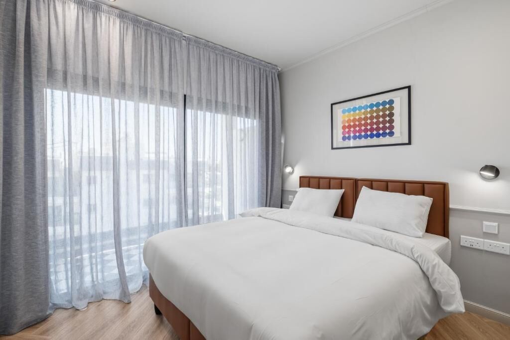 Une chambre d’hôtel avec un grand lit, une fenêtre avec des rideaux légers, une œuvre d’art accrochée au mur et une table de chevet avec une lampe.