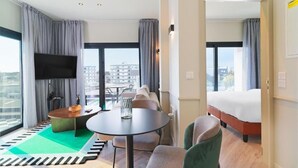 A hotel room with a bed, a TV, a dining table, and a view of the city.