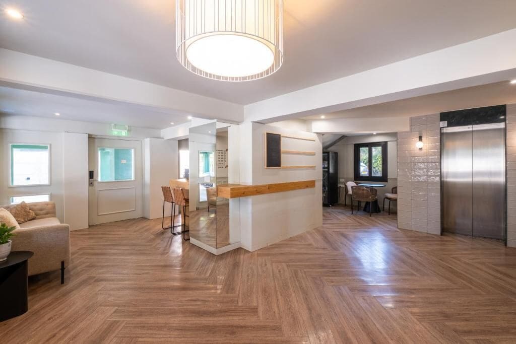A spacious hotel lobby with a central island, wooden flooring, a modern chandelier, and a dining area with chairs.