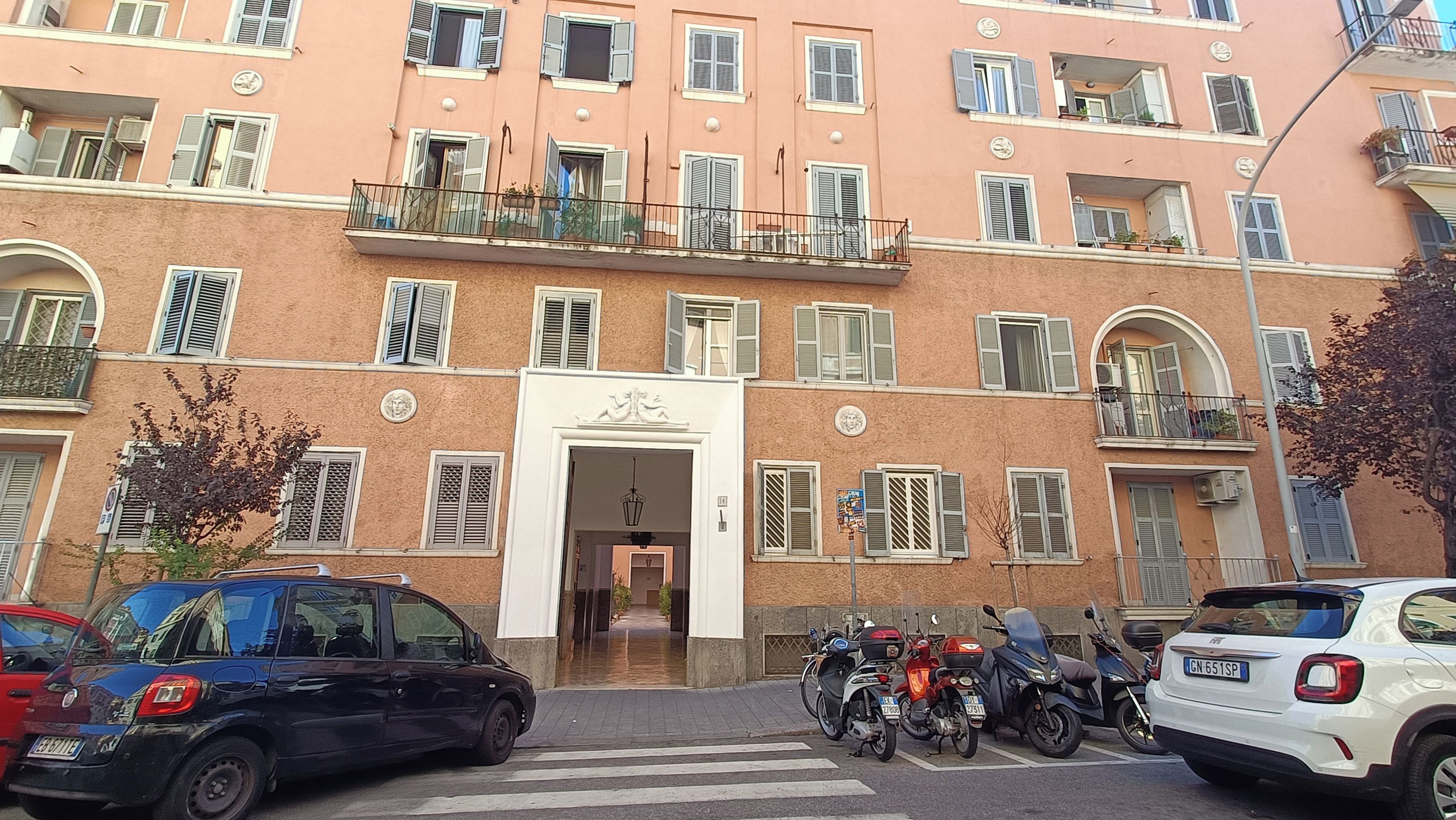 Un immeuble à plusieurs étages doté d’une entrée centrale, de balcons et de véhicules stationnés.