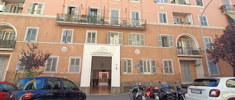 A multi-story building with a central entrance, balconies, and parked vehicles.