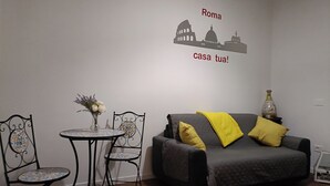 Una stanza con un divano grigio, due cuscini gialli, un tavolino rotondo con un vaso di fiori e un adesivo murale con i monumenti di Roma.