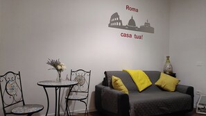 Ein Zimmer mit einem grauen Sofa, zwei gelben Kissen, einem kleinen runden Tisch mit Blumen in einer Vase und einer Wandsticker-Darstellung römischer Sehenswürdigkeiten.