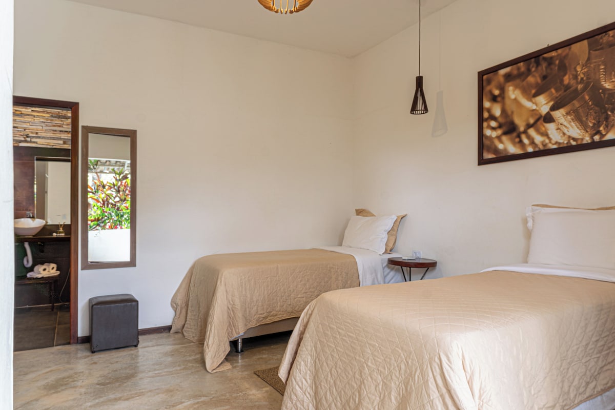 A hotel room with two beds, a small bedside table, a mirror, a window with a view of greenery, and a painting on the wall.