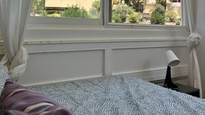 Une chambre avec un lit, une fenêtre avec des rideaux et une vue sur des immeubles et des arbres.