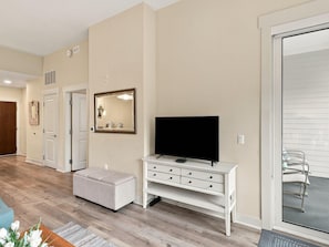 Ein modernes Wohnzimmer mit einem Flachbildfernseher auf einer weißen Konsole, einem großen Spiegel und einer Glastür zum Balkon.