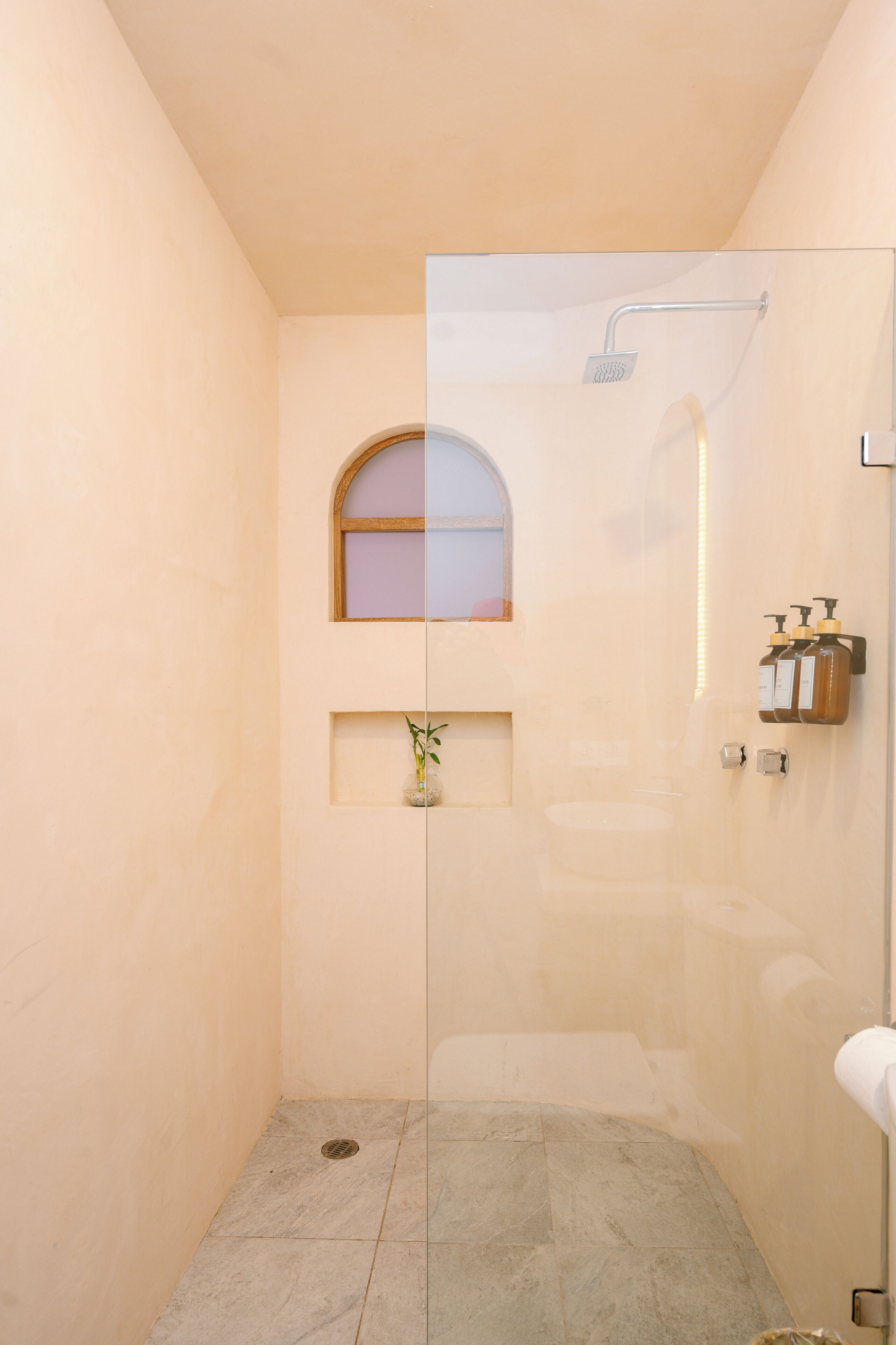 Ducha con cerramiento de vidrio, ventana, un pequeño estante con artículos de aseo y suelo de baldosas.