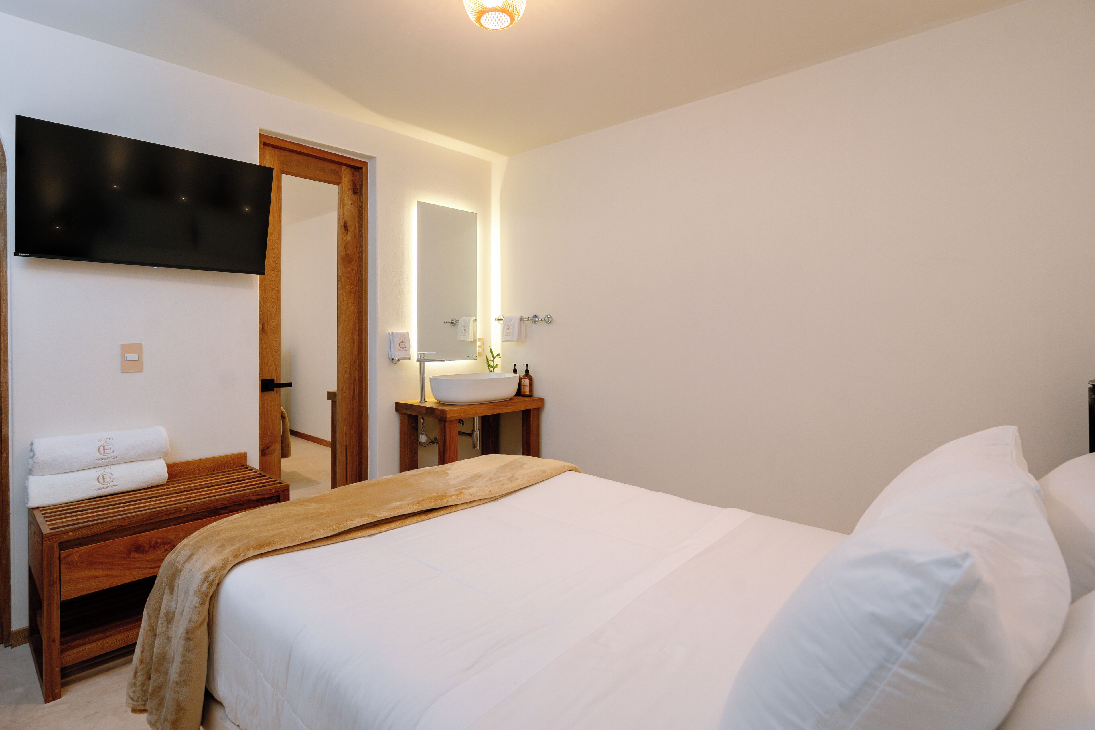 Una habitación de hotel con cama, una mesita de noche de madera, un televisor montado en la pared, un baño con lavamanos y espejo, y una puerta que da a otra habitación.