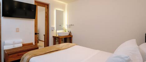 Una habitación de hotel con cama, una mesita de noche de madera, un televisor montado en la pared, un baño con lavamanos y espejo, y una puerta que da a otra habitación.