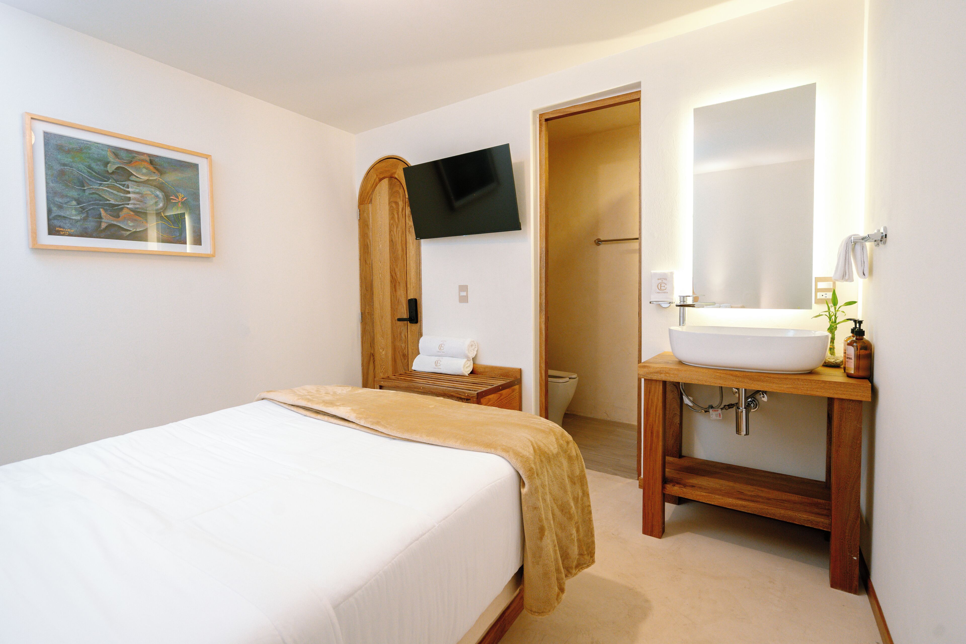 A hotel room with a bed, a wooden nightstand, a television mounted on the wall, a bathroom with a sink and mirror, and a painting on the wall.