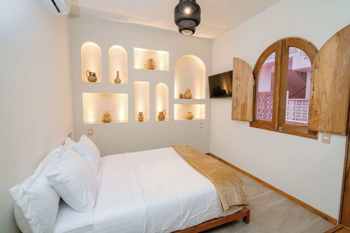 A bedroom with a bed, wall-mounted shelves with pottery, a wooden door, and a ceiling light fixture.