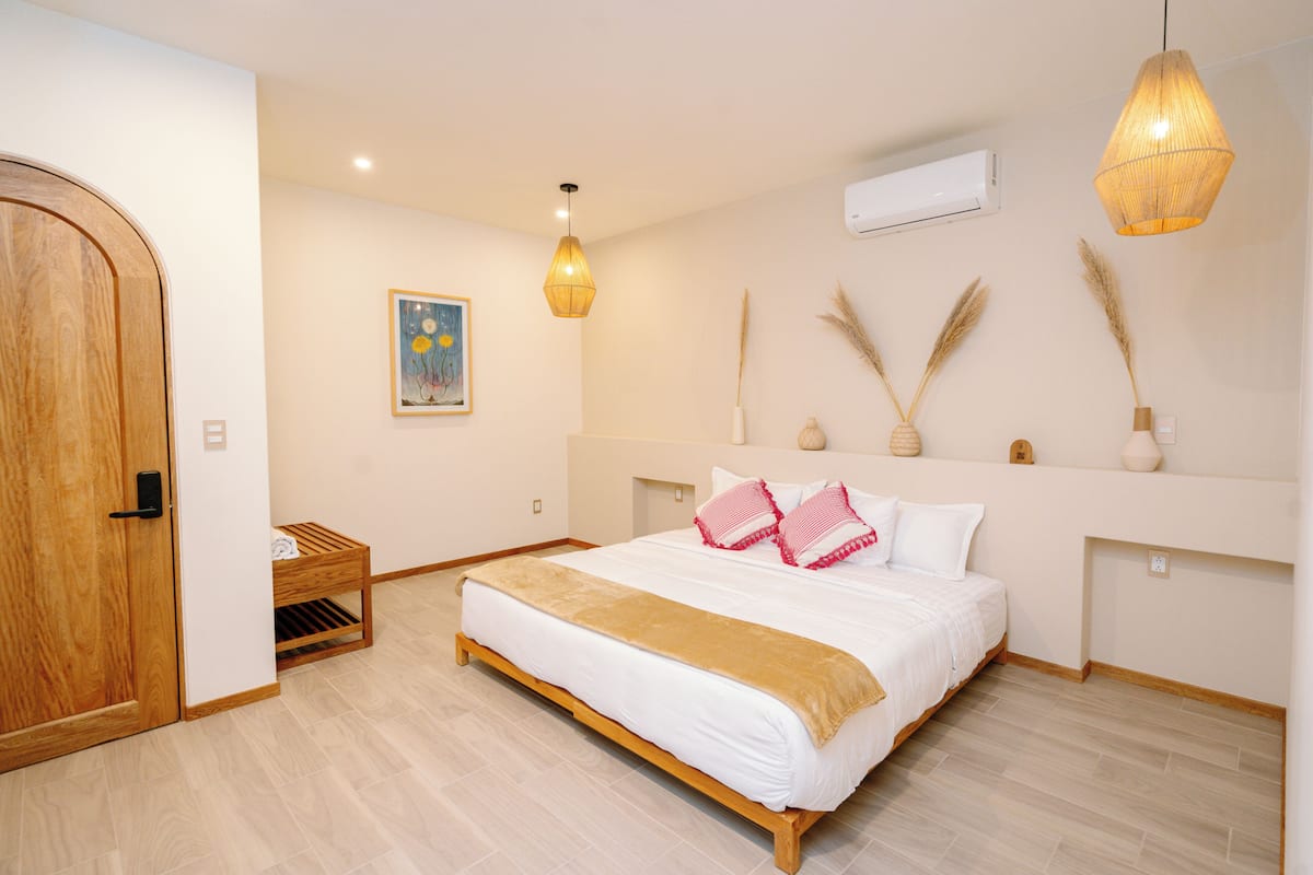 A bedroom with a bed, wooden door, bedside table, wall-mounted air conditioner, pendant lights, and a framed picture.