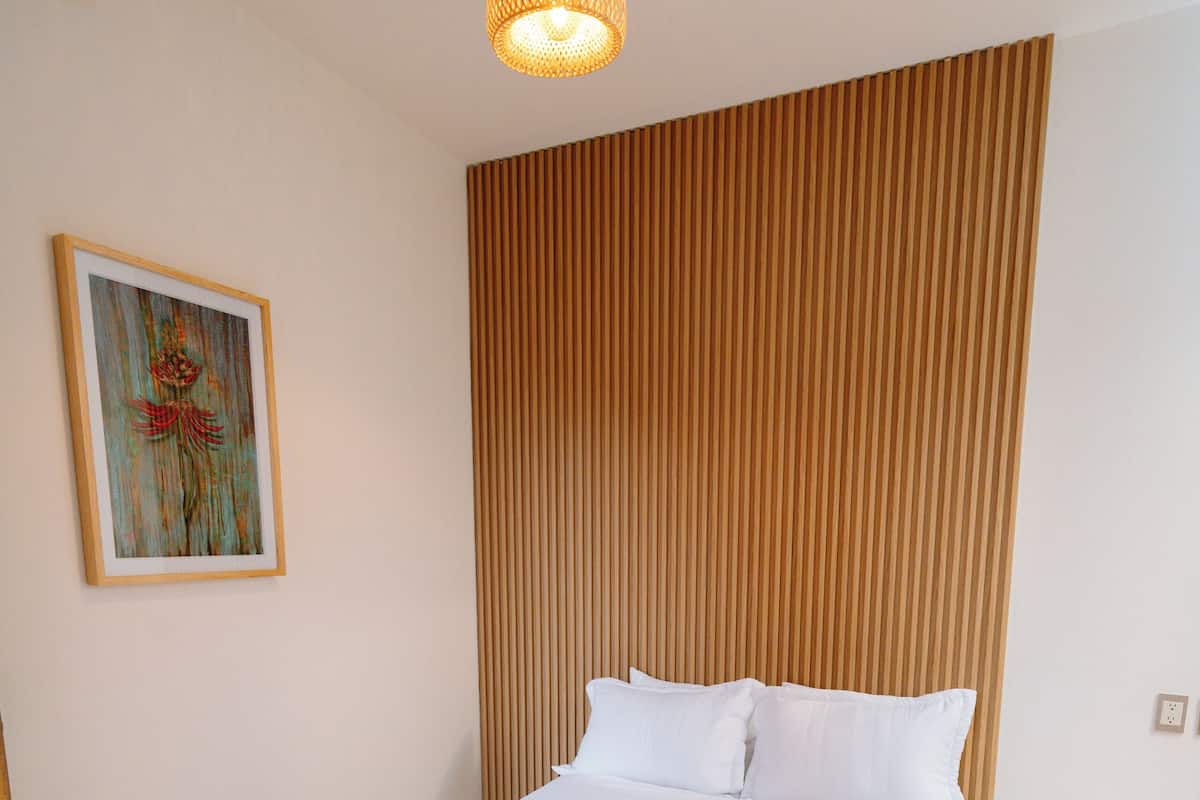 A neatly made bed with white linens and a brown throw, a wooden headboard, a hanging light fixture, and a framed picture on the wall.