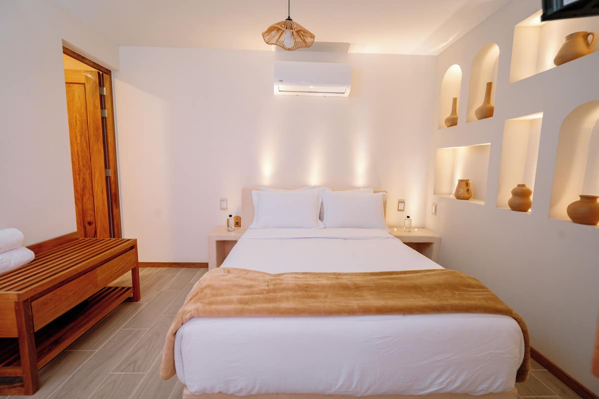 A bedroom with a wooden bench, a bed with white bedding, a brown throw blanket, a bedside table with a lamp, and wall-mounted pottery.