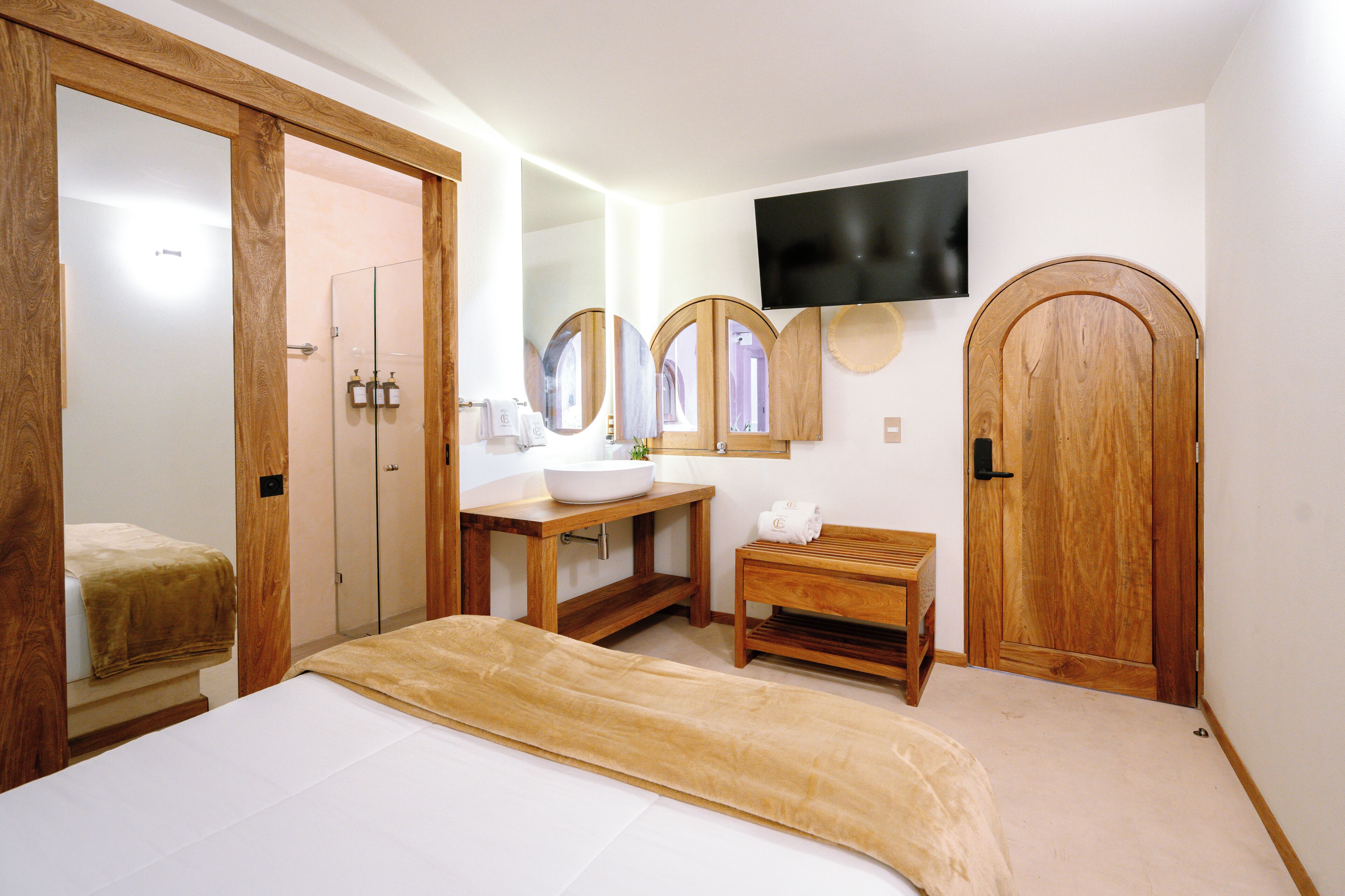 A hotel room with a bed, a wooden bench, a vanity with a mirror, a television mounted on the wall, and a wooden door.