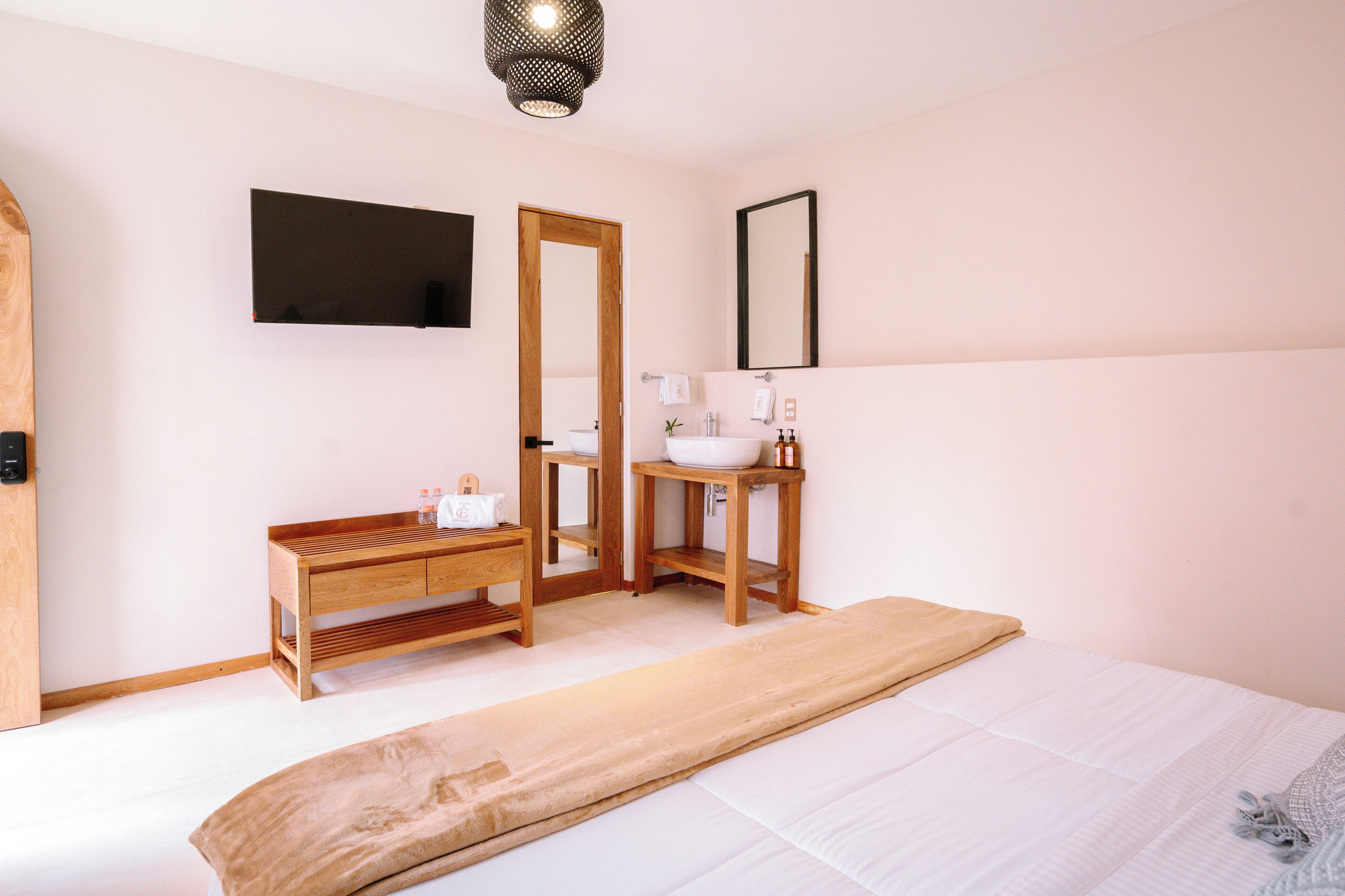 Un dormitorio con cama, mesitas de noche de madera, un espejo grande, televisor montado en la pared y un baño con lavabo e inodoro.
