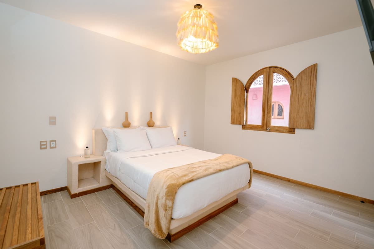 A bedroom with a bed, bedside tables, a wooden bench, a hanging light fixture, and a window with wooden shutters.