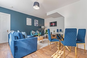 A modern living room with a blue sofa, matching chairs, a dining table, and a TV mounted on the wall. - Stylish 2BR House close to Camden Town Station (London)