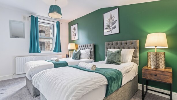 Una cama doble con cabecera gris, una mesita de noche con lámpara y una ventana con cortinas color aguamarina.