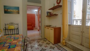 Ein Zimmer mit einem Bett, einem Tisch mit farbigem Tischlein, einer kleinen Kommode, einem Bücherregal, einer Uhr und einem Fenster mit Blick auf Gebäude.