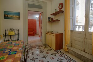 Ein Zimmer mit einem Bett, einem Tisch mit farbigem Tischlein, einer kleinen Kommode, einem Bücherregal, einer Uhr und einem Fenster mit Blick auf Gebäude.
