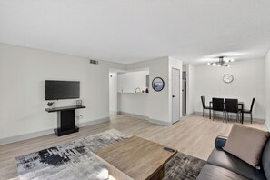 A modern living room with a flat-screen TV, a coffee table, a sofa, a dining area with chairs, and a clock on the wall. - Contemporary 2BR apartment with workspace (Lenexa)