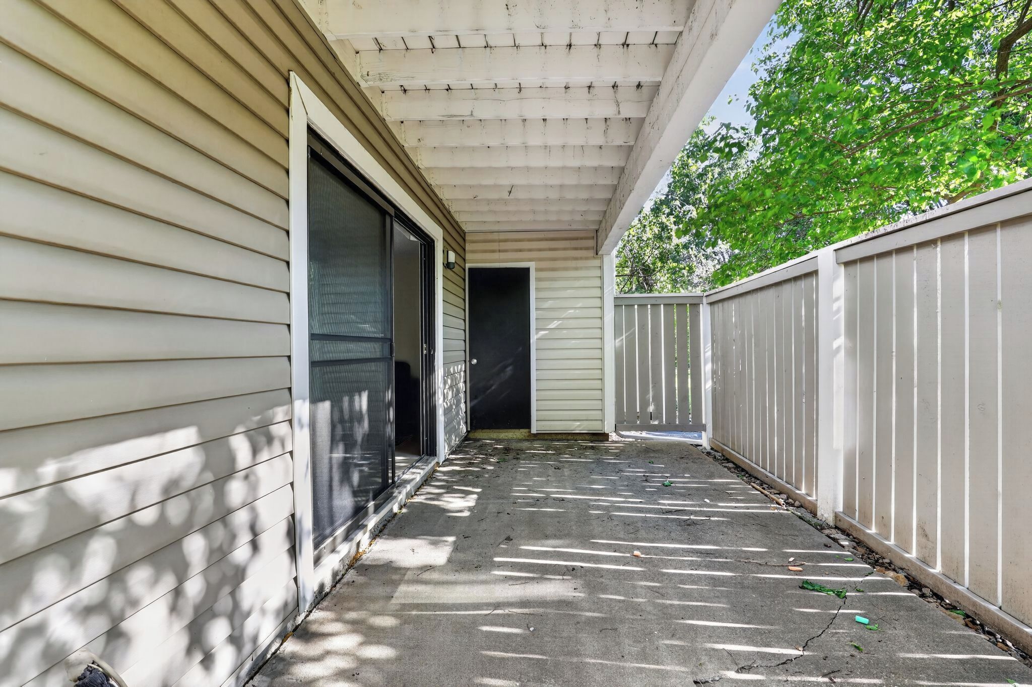 A covered outdoor area with a concrete floor, a white fence, and a building with a garage door.