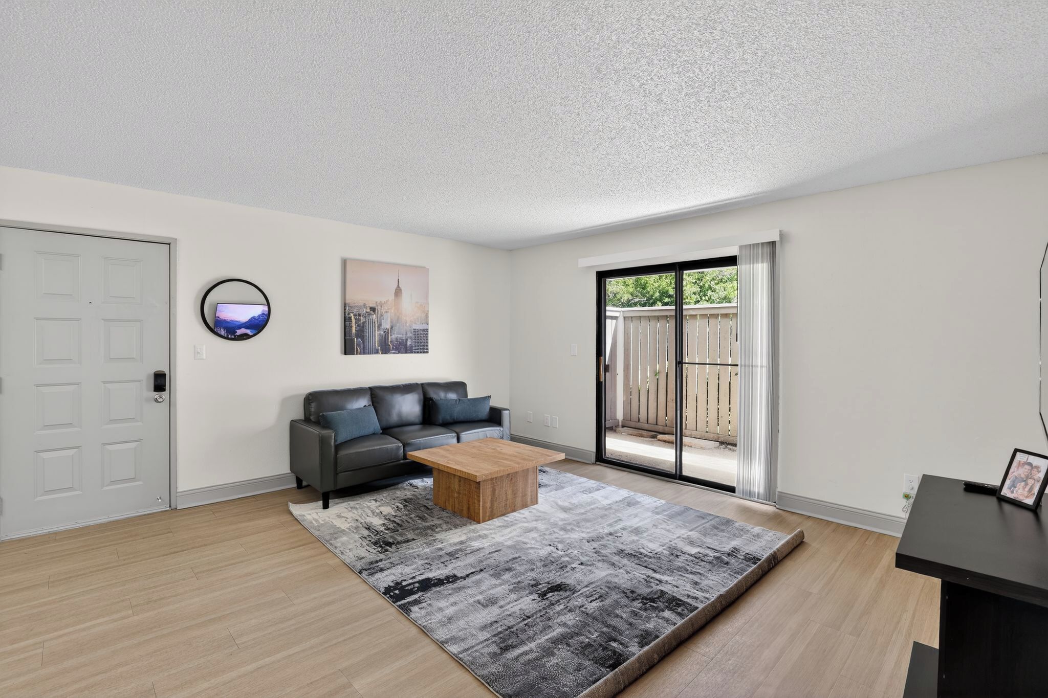 A modern living room with a gray sofa, a wooden coffee table, a gray rug, a cityscape painting, and a sliding glass door leading to a patio.