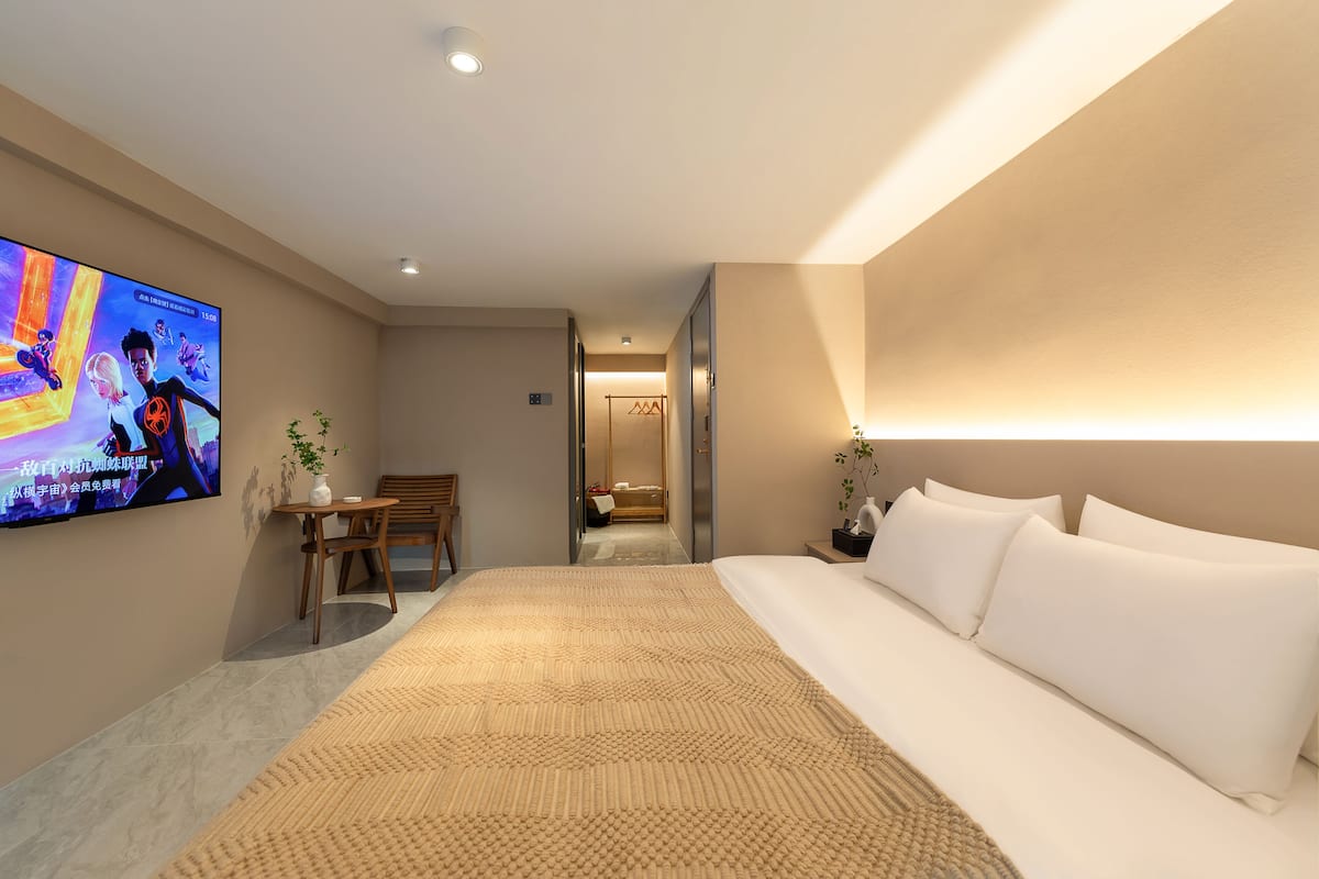 A hotel room with a large bed, a television on the wall, a desk with a chair, and a bathroom visible through an open door.