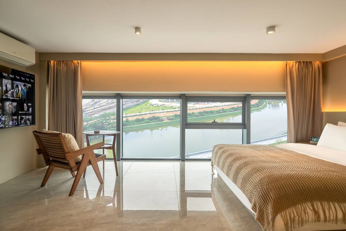 A hotel room with a large window, a bed, a chair, a small table, and a wall-mounted TV.