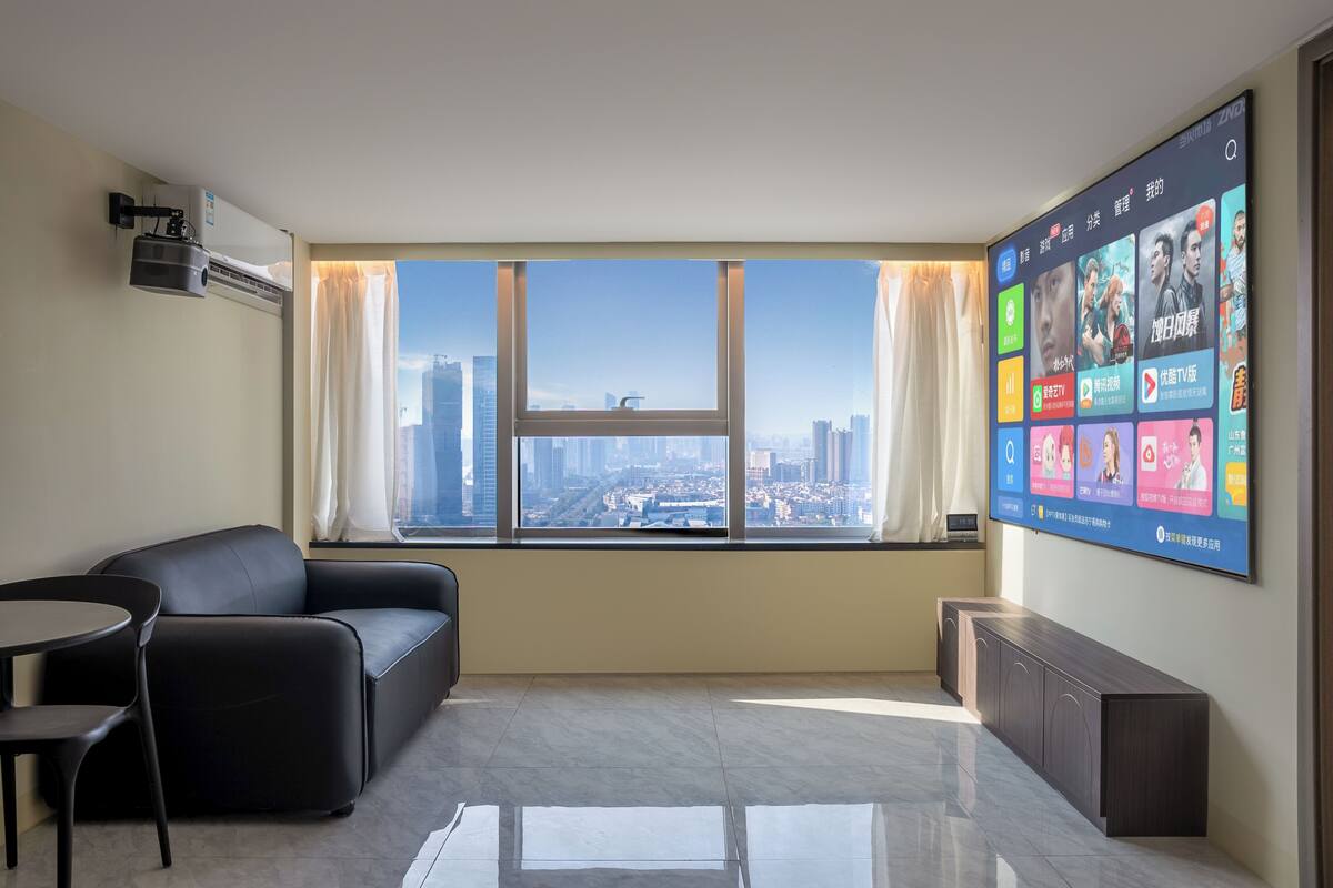 A modern hotel room with a large window, a black leather sofa, a small table, and a flat-screen TV mounted on the wall.