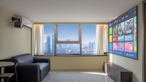 A modern hotel room with a large window, a black leather sofa, a small table, and a flat-screen TV mounted on the wall.