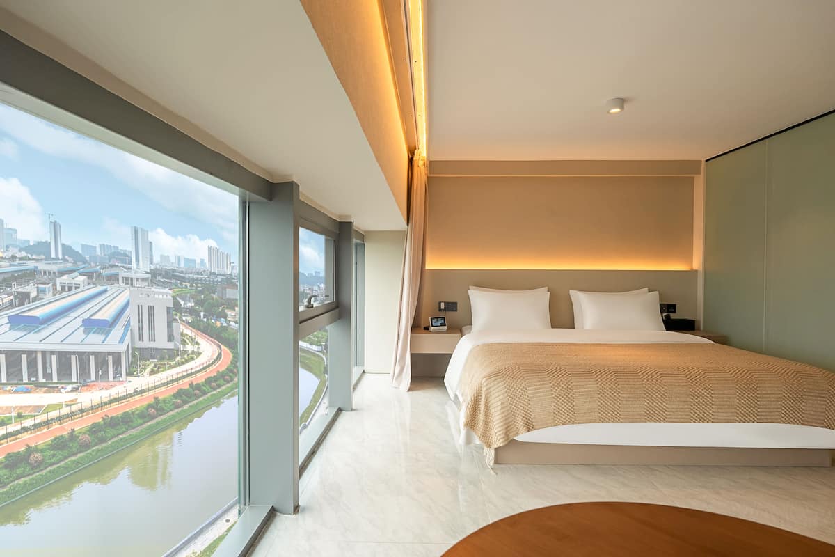 A modern hotel room with a large window, a bed, a bedside table, and a view of a cityscape.