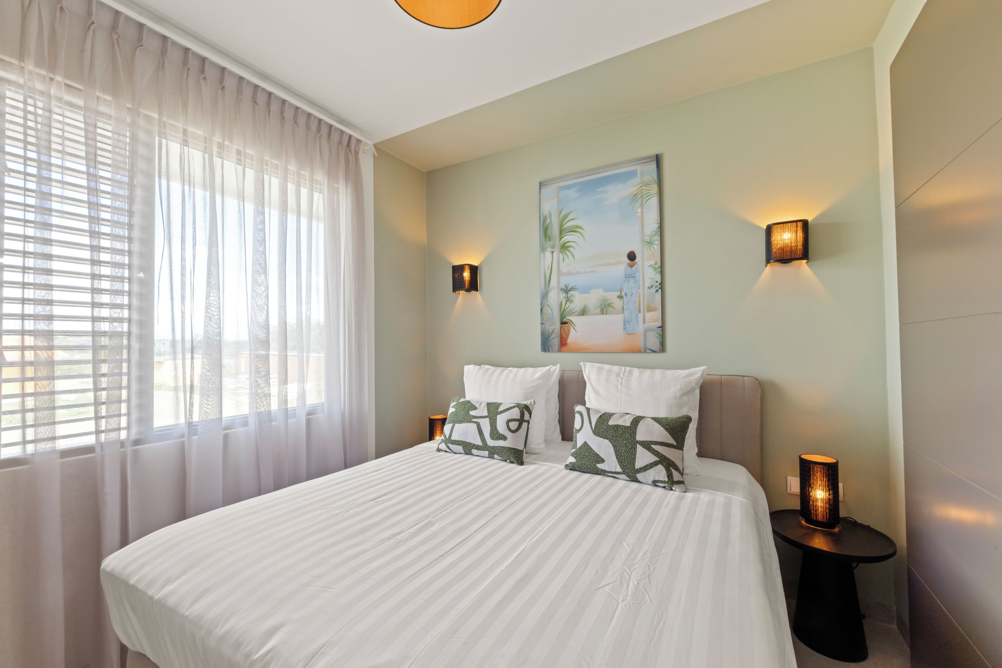 A bedroom with a bed, two bedside tables, a painting, and a window with curtains.