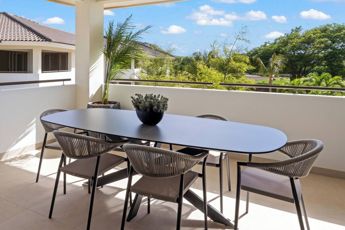 Zona pranzo con tavolo e sedie, una ventola a soffitto e vista su vegetazione ed edifici.