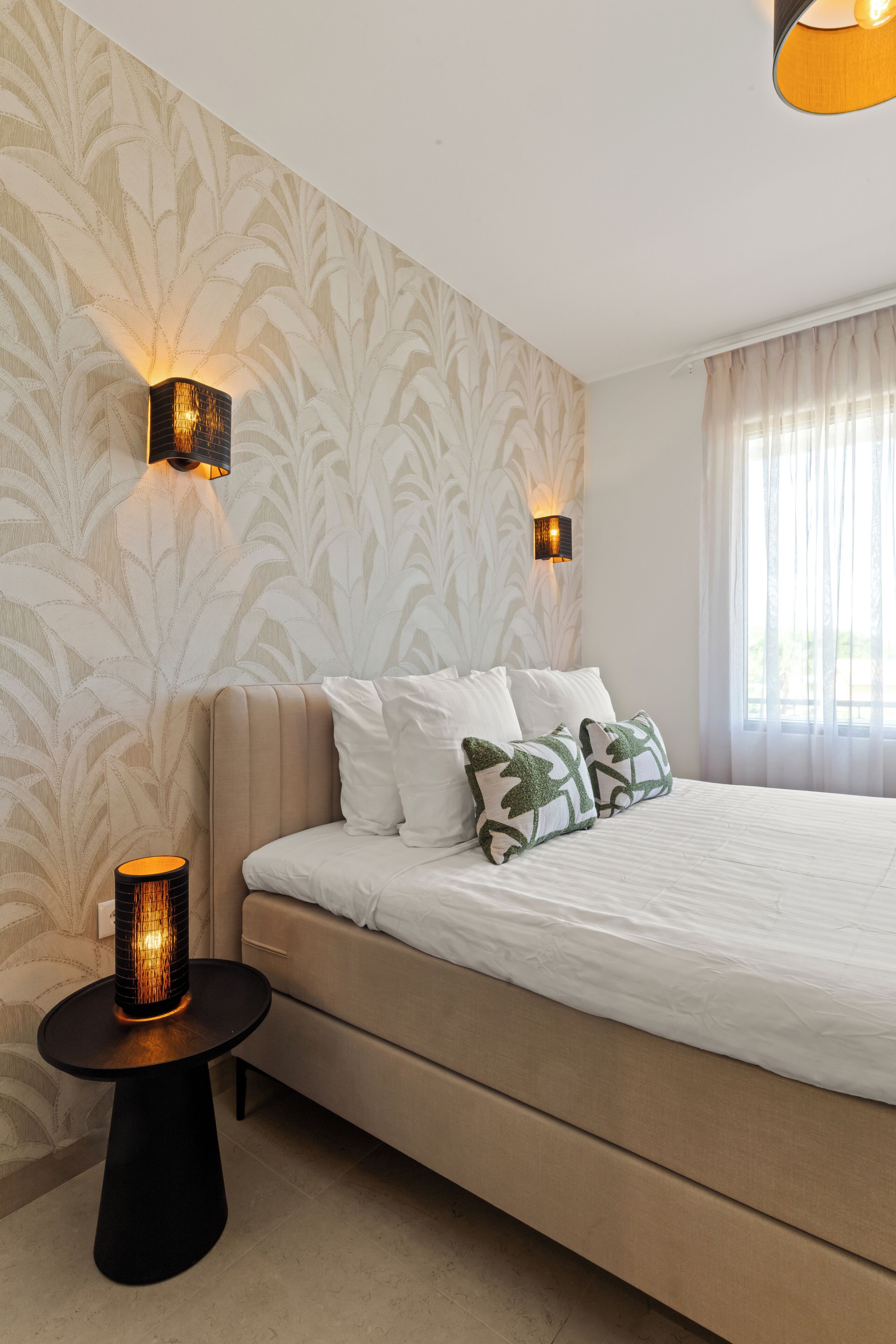 A bedroom with a bed, two wall-mounted lamps, a round table with a lamp, and a patterned wallpaper.