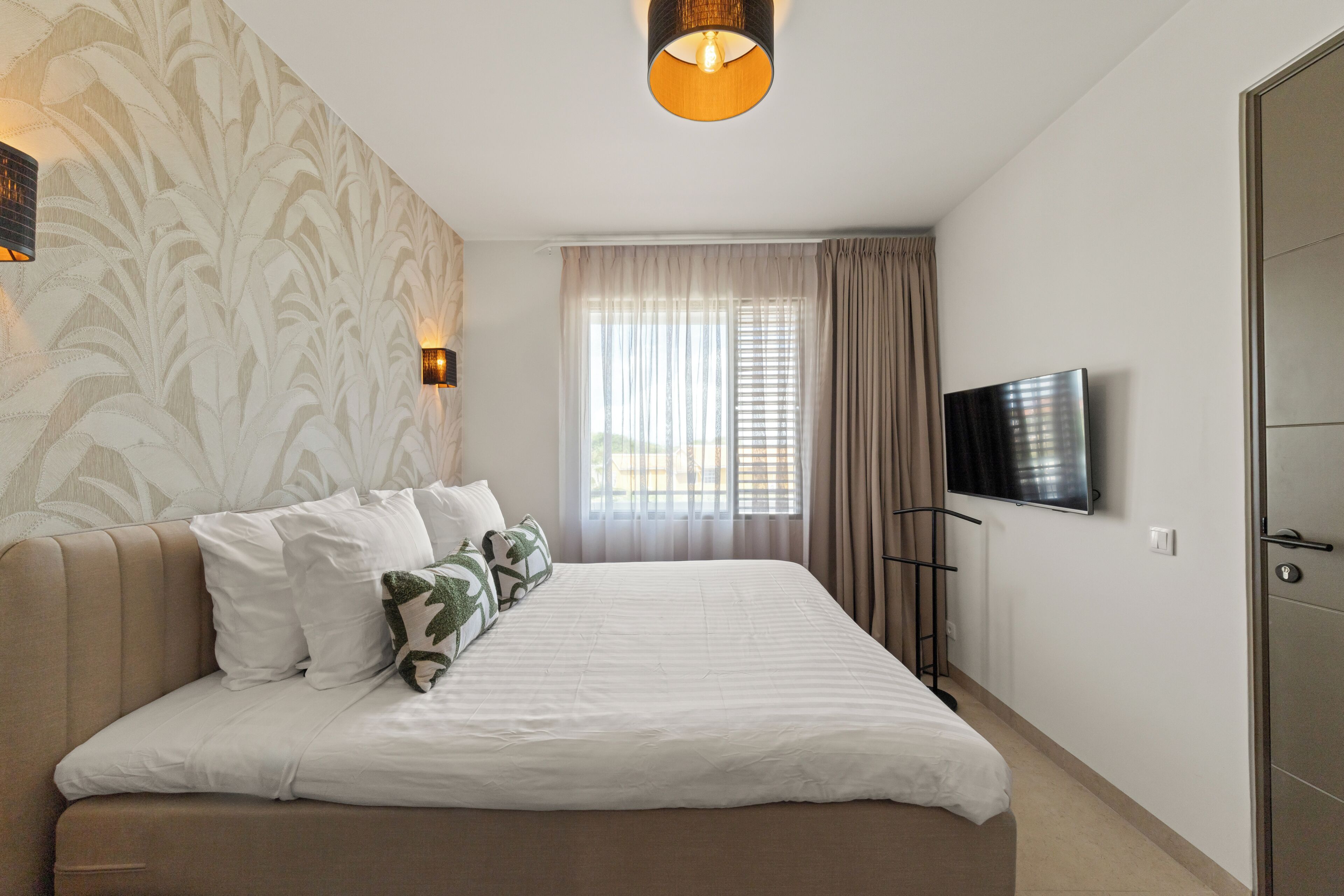 Une chambre d’hôtel avec un lit, une télévision, une fenêtre avec des rideaux et un luminaire fixé au mur.