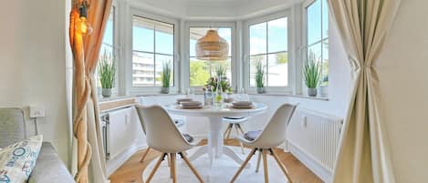 A corner room with a round table, two chairs, a hanging light fixture, and large windows.