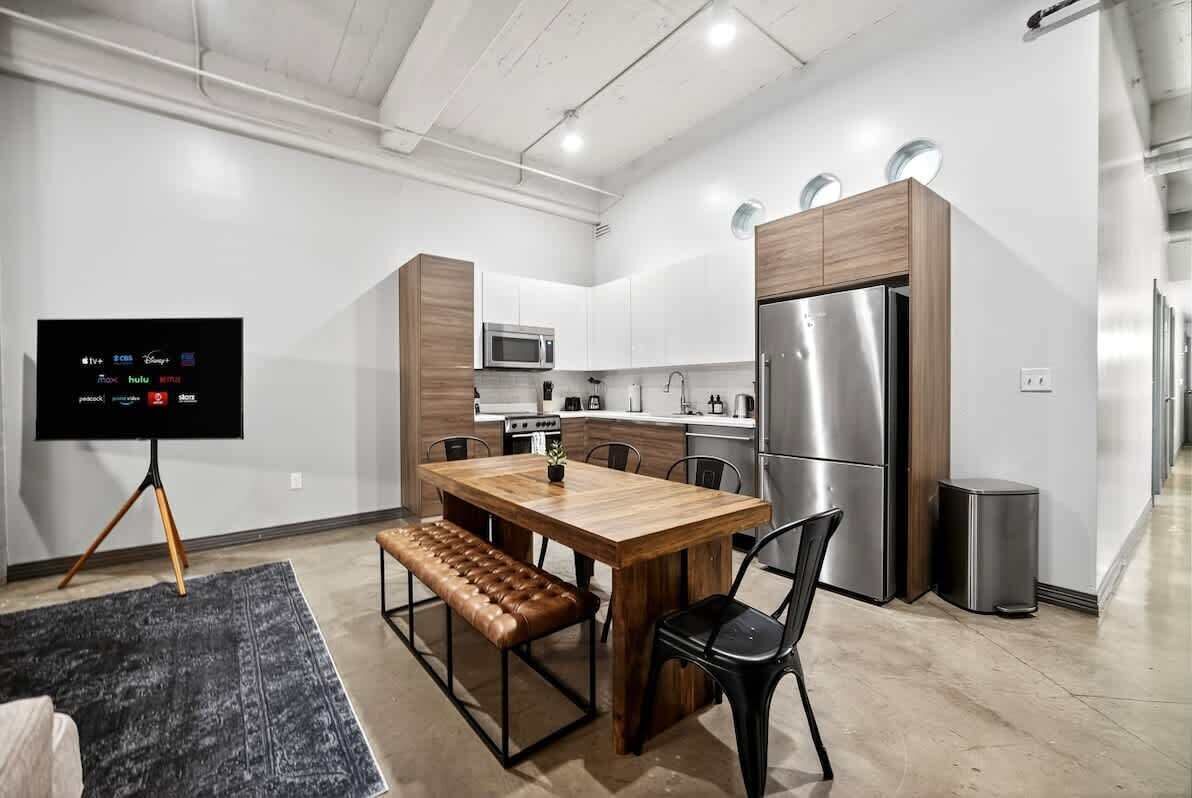 Una cocina moderna con una mesa de comedor de madera, sillas negras, un banco, un televisor sobre un trípode y electrodomésticos de acero inoxidable.