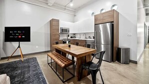 Une cuisine moderne avec une table à manger en bois, des chaises noires, un banc, un téléviseur posé sur un trépied et des appareils électroménagers en acier inoxydable.