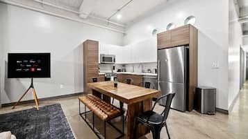 Una cocina moderna con una mesa de comedor de madera, sillas negras, un banco, un televisor sobre un trípode y electrodomésticos de acero inoxidable.
