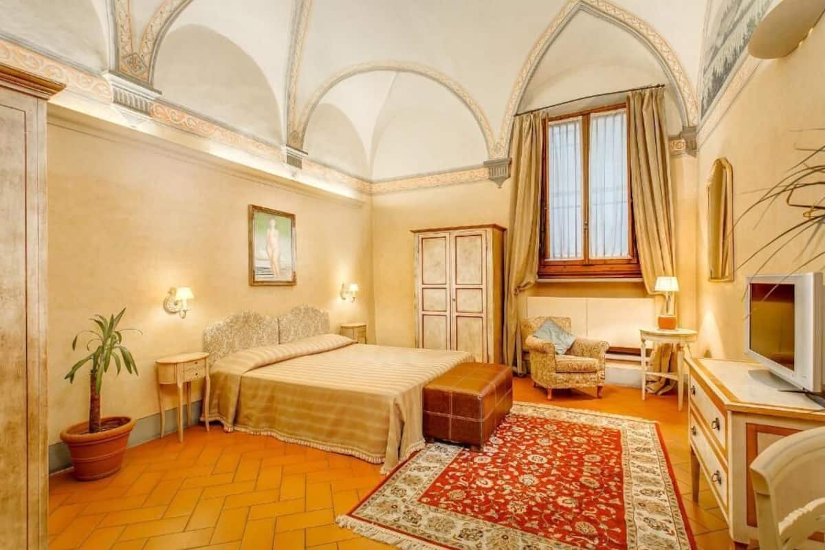 Un dormitorio con una cama, una silla, un televisor, una planta en maceta y una ventana con cortinas.