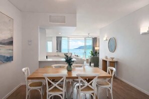 A dining area with a wooden table and white chairs, a view of the sea through large windows, and a modern interior design. - Luxury 3BR Coral Coast Home w/ Oceanfront (Cairns City)