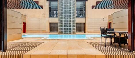 Uma área de piscina com um pilar revestido em azulejos de vidro, piso ladrilhado e uma mesa com cadeiras.