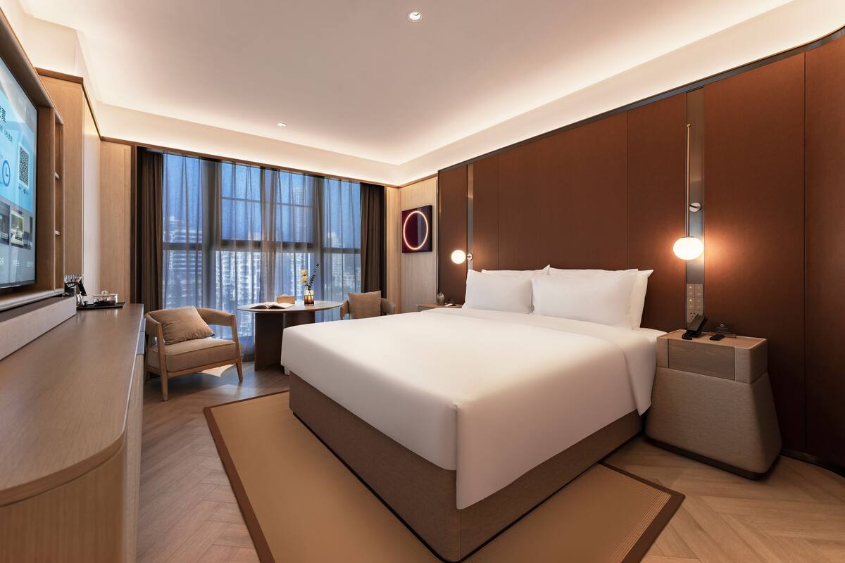 A modern hotel room with a large bed, a desk, a chair, and a large window.