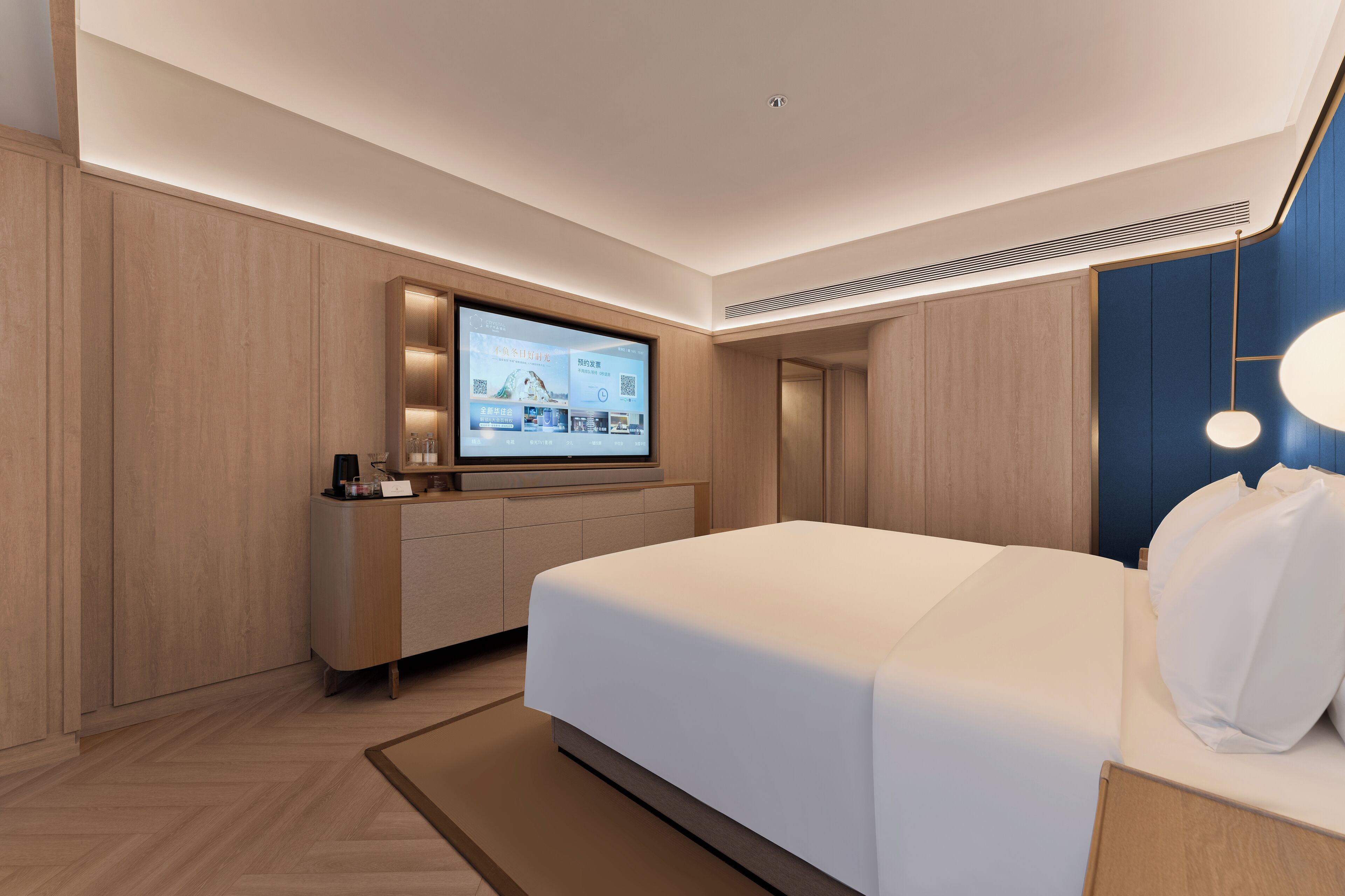A modern hotel room with a large bed, a built-in TV, a desk with a computer, and a ceiling vent.